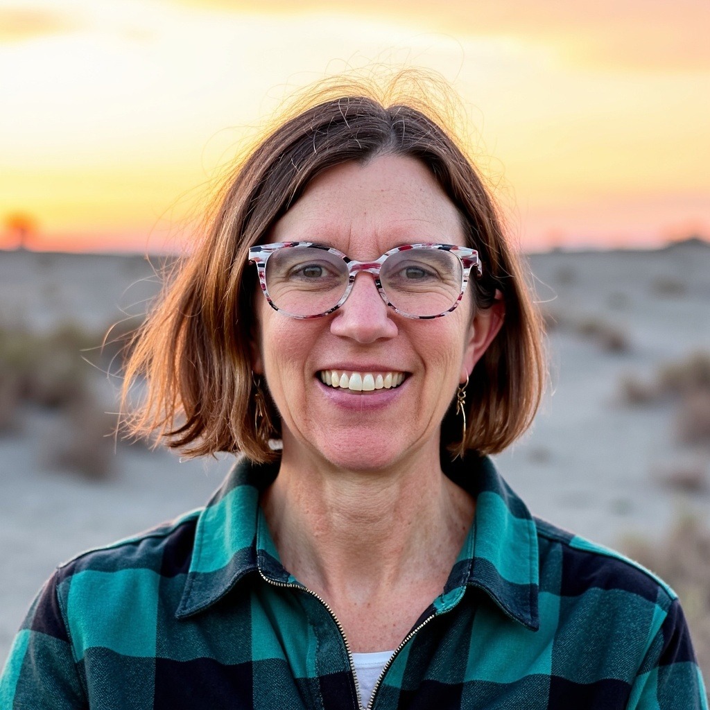 Woman with short brown hair, wearing colorful glasses and a green plaid shirt, smiles against a sunset backdrop in a desert setting.